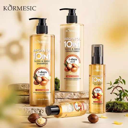 Four Körmesic hair care products arranged on stone blocks with whole and halved argan nuts and small greenery props against a warm beige background. The set includes shampoo, conditioner, a small spray bottle, and a tube, all in transparent bottles with golden liquid and black pumps/caps.