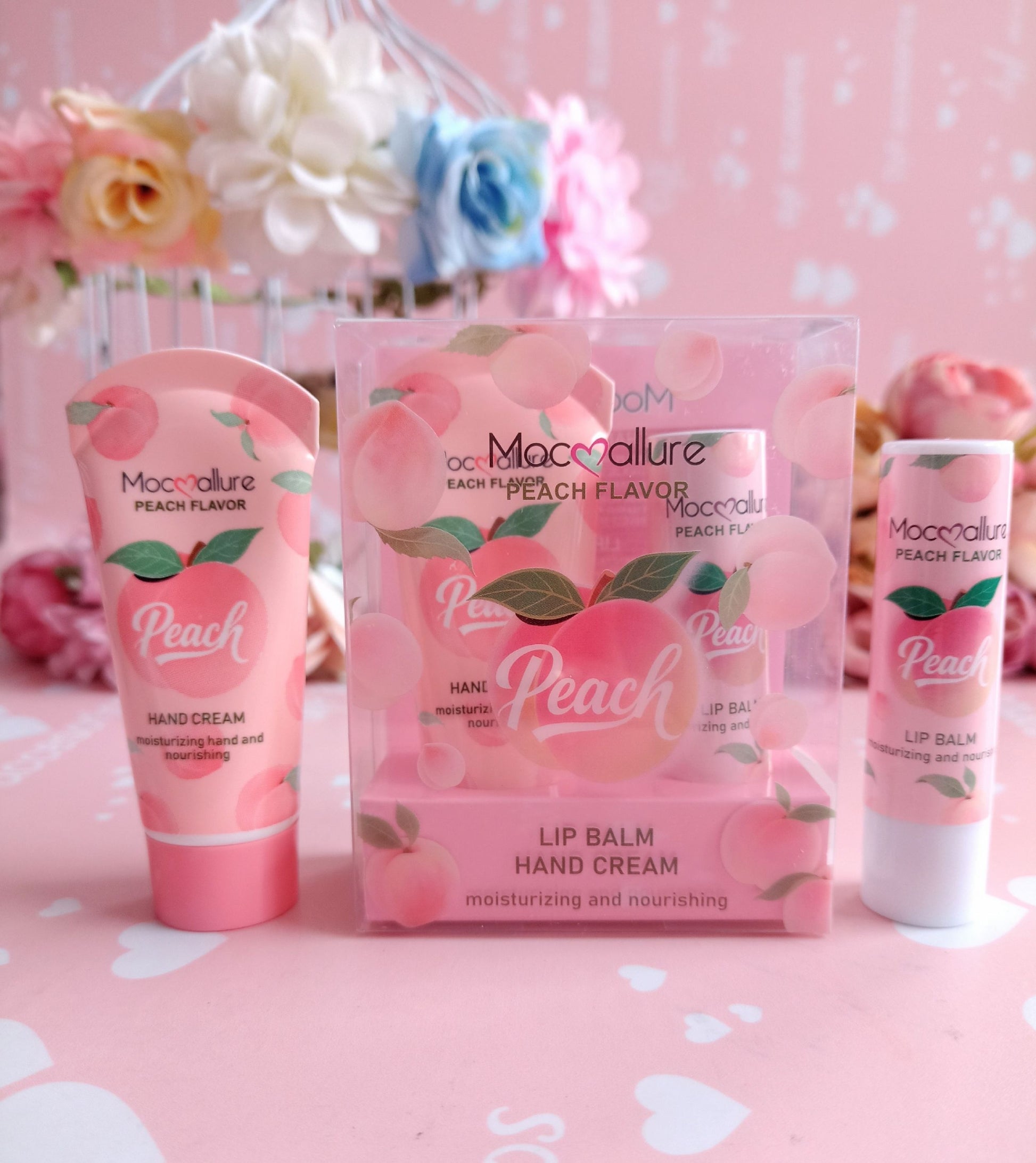 A set of Macallure Peach Flavor hand cream and lip balm products arranged on a pink surface with a floral backdrop. The set includes a hand cream tube, a clear box of hand cream, and a lip balm tube.