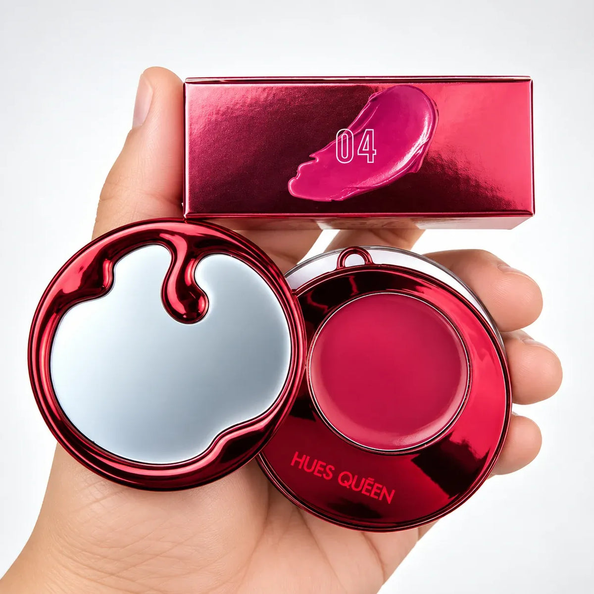 A hand holds a round red makeup compact with a mirror lid and a pink cream product, alongside a matching red box marked '04' on a white background.