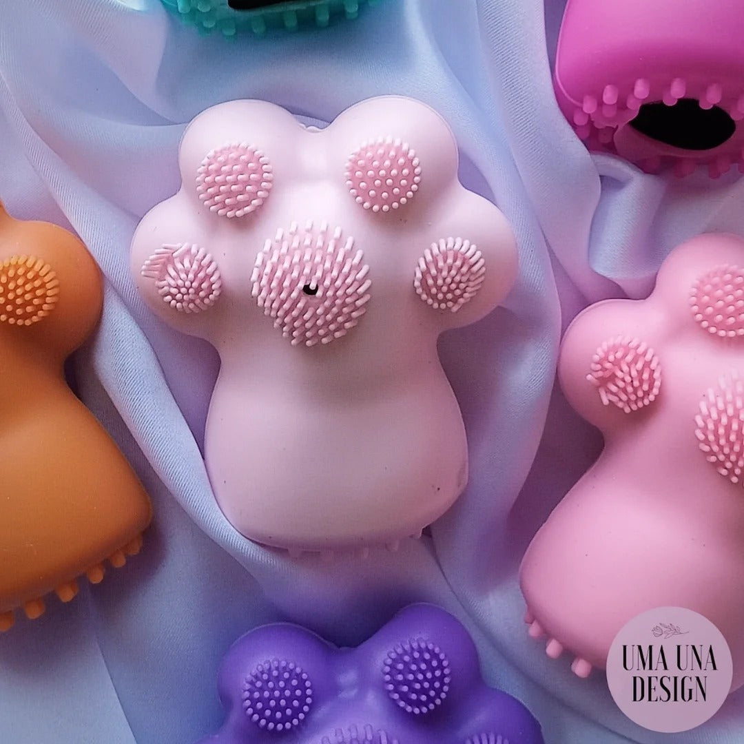 A set of pastel-colored silicone pet massage toys with textured nubs, arranged on a draped lavender fabric background. The visible toys are pink, orange, purple, and magenta.