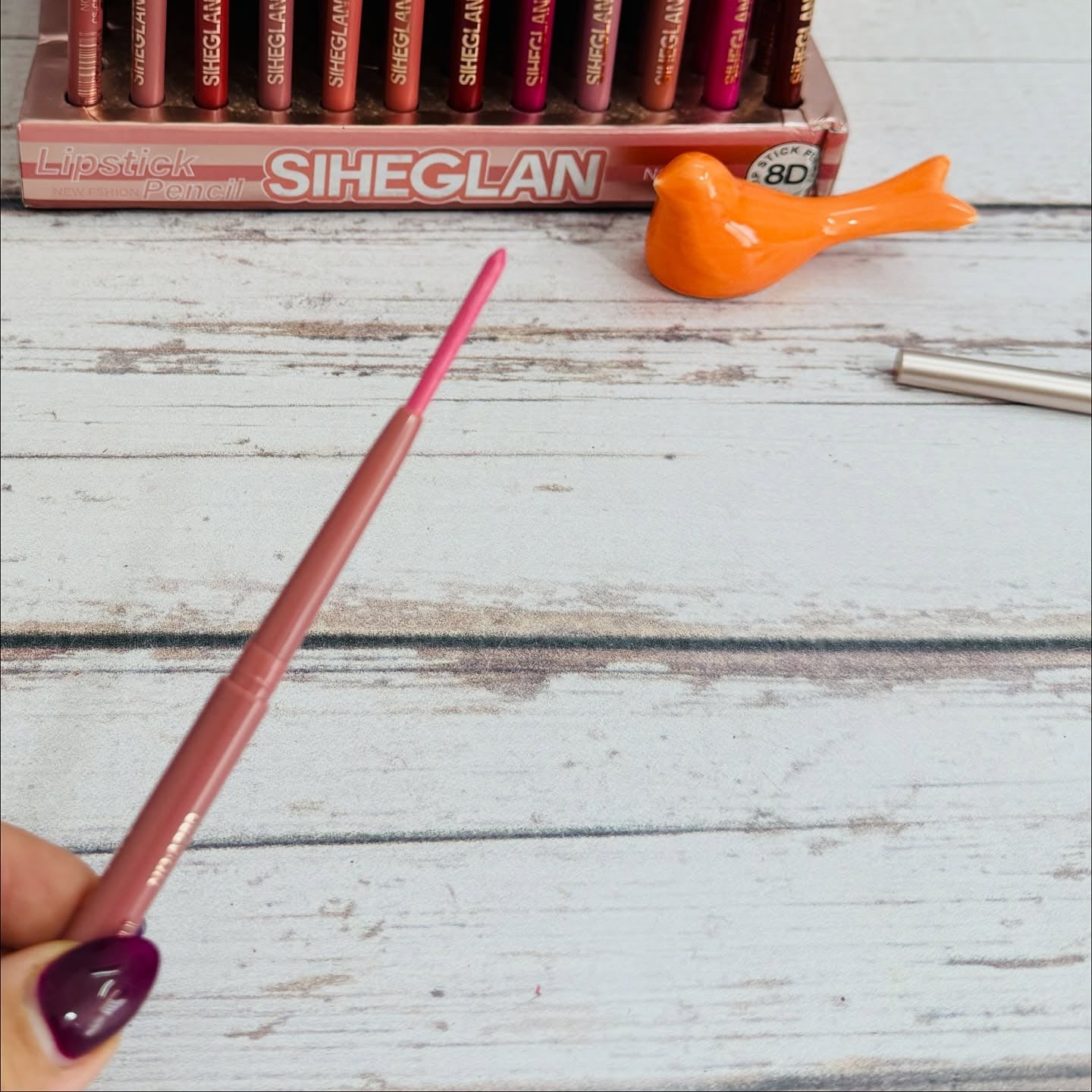 A hand with dark purple nail polish holds a pink lip liner pencil in the foreground, with a box of Siheglan lipsticks and an orange bird-shaped cosmetic accessory blurred in the background on a white wooden surface.