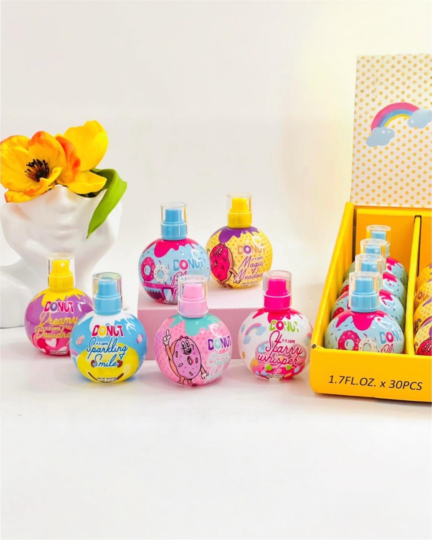 A set of colorful round perfume atomizer bottles with spray caps, displayed alongside their yellow retail box on a white surface. A white vase with yellow flowers is visible in the background on the left.