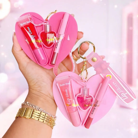 A hand holds two pink heart-shaped lip gloss keychains with gold rings and charms. Each keychain contains a pink heart-shaped case with a lip gloss tube, a small pink heart-shaped lip gloss bottle, and a pink pen.