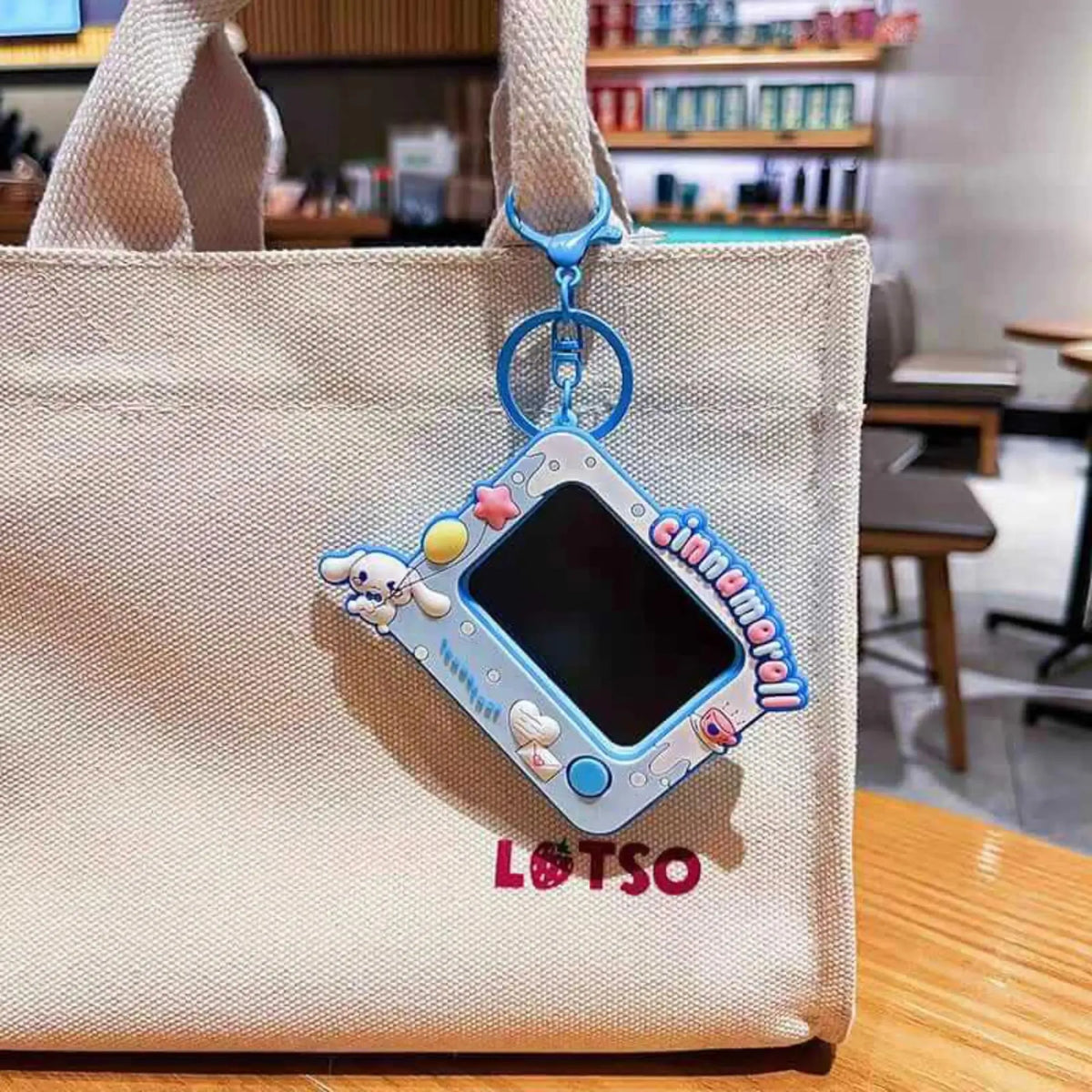 A light blue and white Cinnamoroll-themed handheld toy camera charm with a strap, hanging from a beige canvas tote bag in a cafe setting.