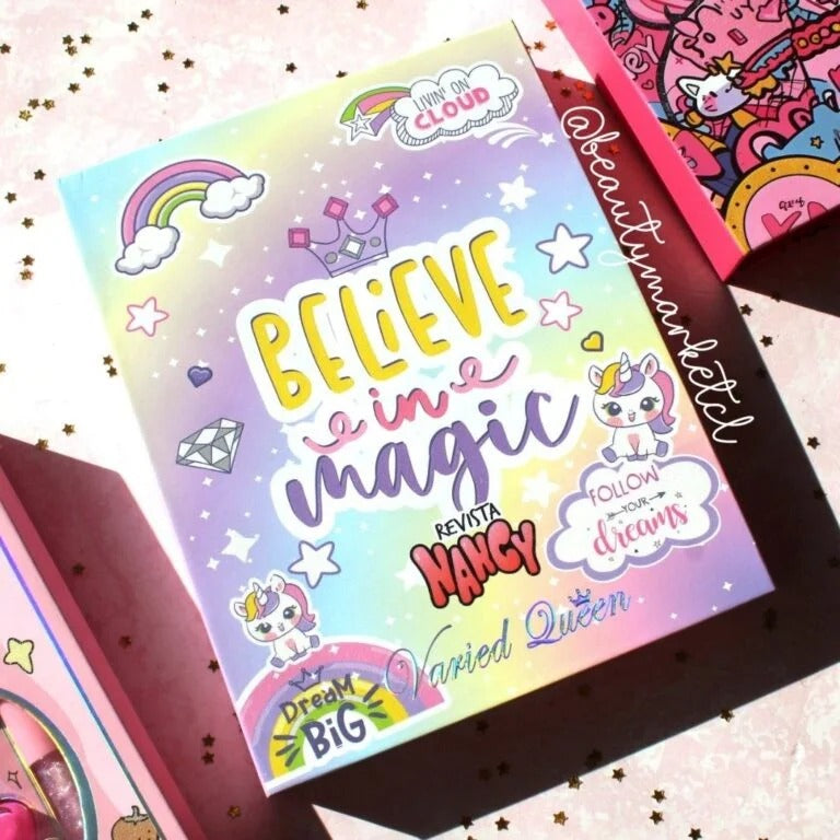 A colorful illustrated card or booklet cover reading 'BELIEVE in magic' with unicorn, rainbow, cloud, and star graphics, photographed on a pink glittery surface with other similarly styled items partially visible around it.
