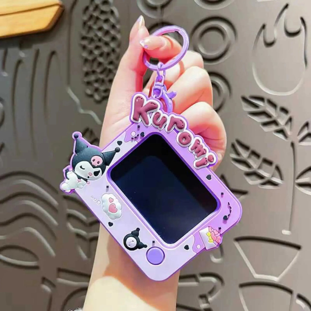 A hand holding a purple Kuromi-themed Tamagotchi-style toy with a screen and cartoon character details, set against a patterned gray background.