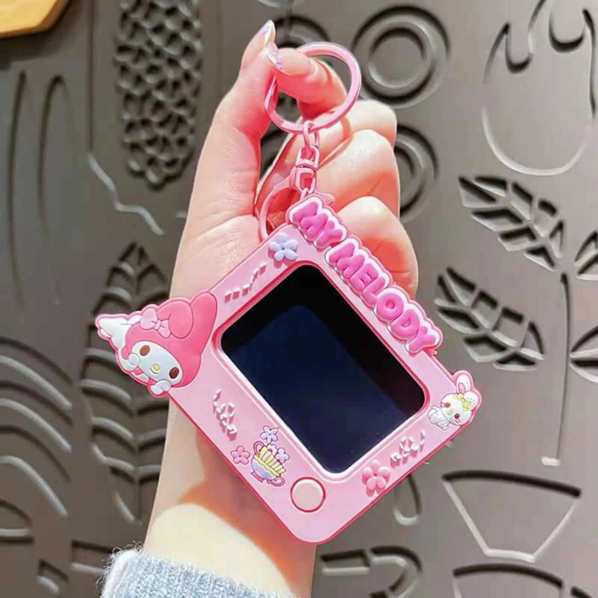 A hand holding a pink My Melody digital toy camera keychain against a gray patterned background.