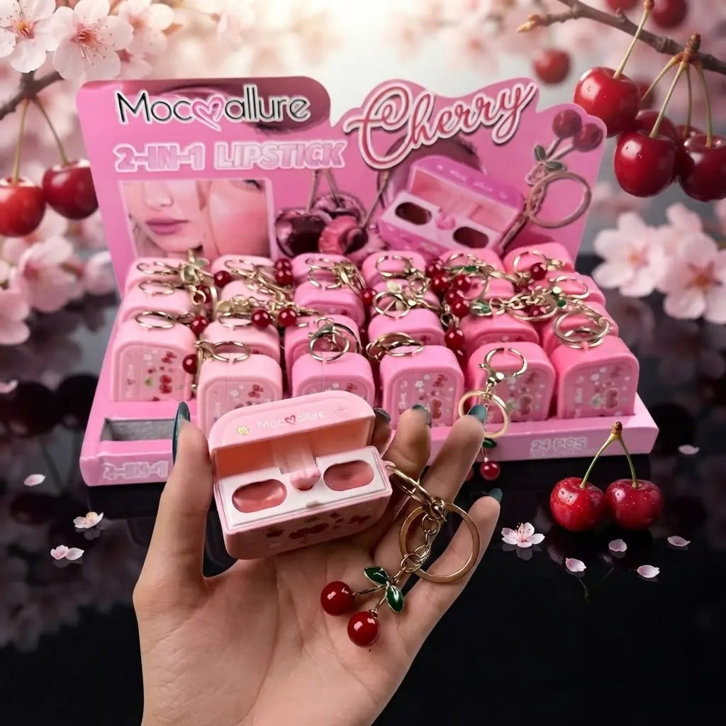 A hand holds a pink cherry-themed lipstick case with a cherry keychain in front of a retail display of matching pink lipstick cases and keychains, with cherry branches and pink blossoms in the background.