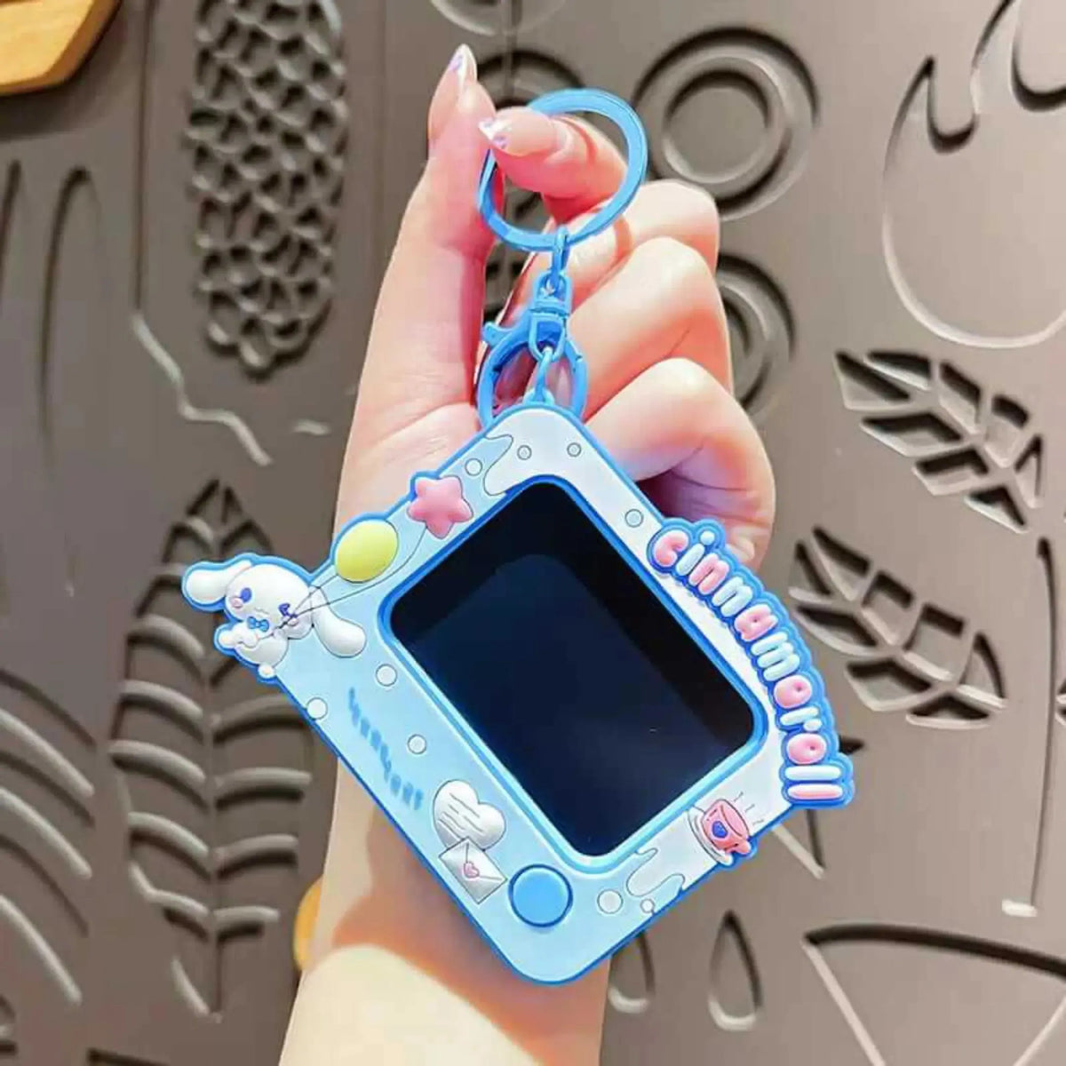 A hand holding a light blue Cinnamoroll-themed electronic toy keychain against a gray patterned metal background.