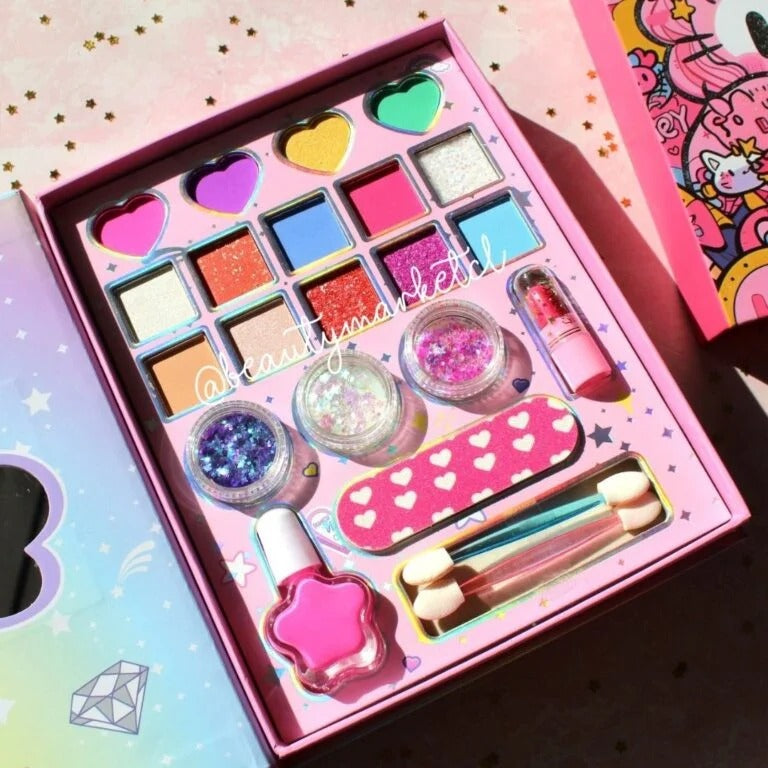 A pink makeup kit displayed in an open box, featuring heart-shaped eyeshadow pans, glitter jars, a pink nail polish bottle, dual-ended applicators, and a heart-patterned nail file. The kit is photographed on a pastel pink glittery surface with a Hello Kitty box partially visible at the top right.