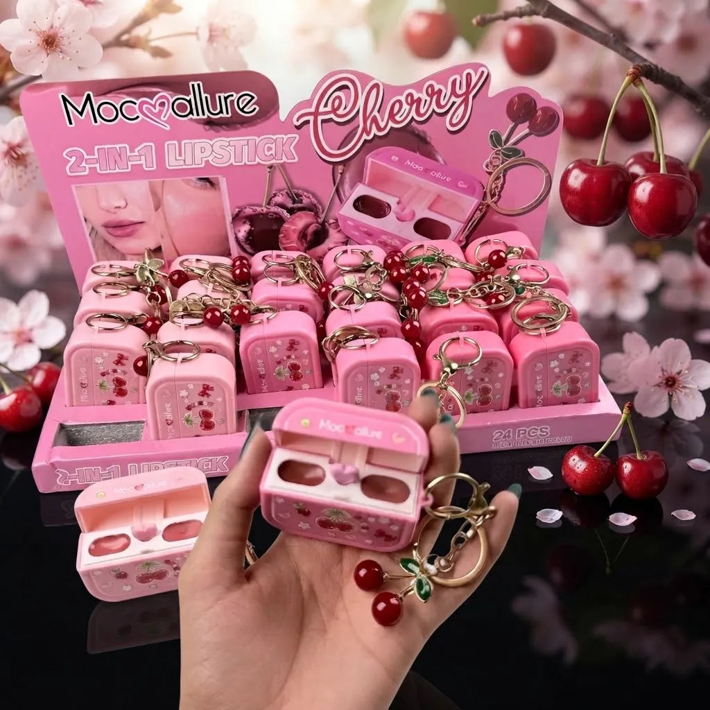 A hand holds a pink cherry-themed lipstick case keychain in front of a retail display of matching pink lipstick cases and keychains. The display features cherry illustrations, the text 'Moc•allure', and 'Cherry', with red cherries and pink blossoms in the background.