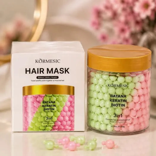 Körmesic Hair Mask jar and packaging box on a light surface with pink flowers blurred in the background. The clear jar contains pink and green round hair mask beads with a gold lid, next to a white box showing the same color scheme.