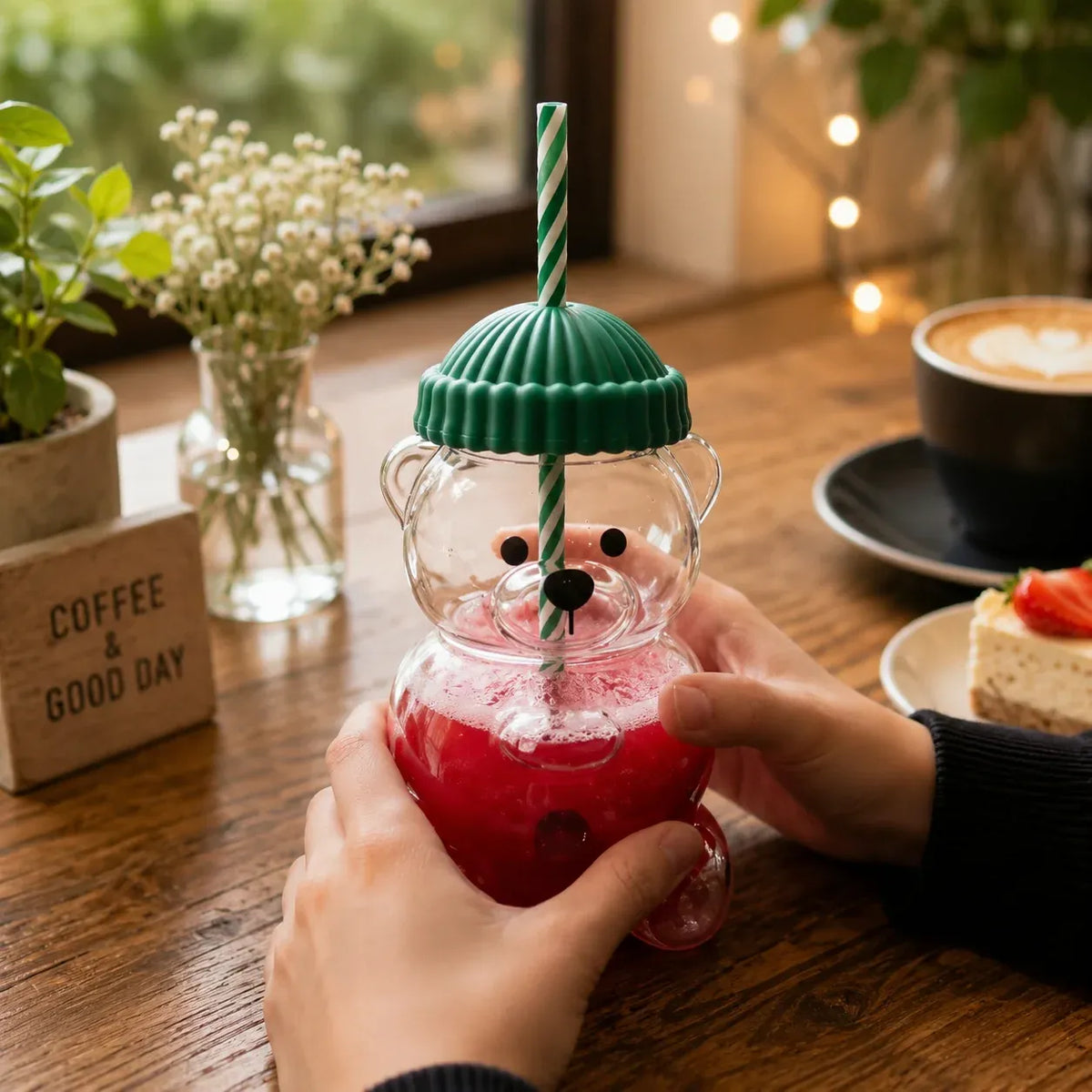 Person holding a bear-shaped glass with a red drink and green lid on a wooden table with a cup of coffee and cake.