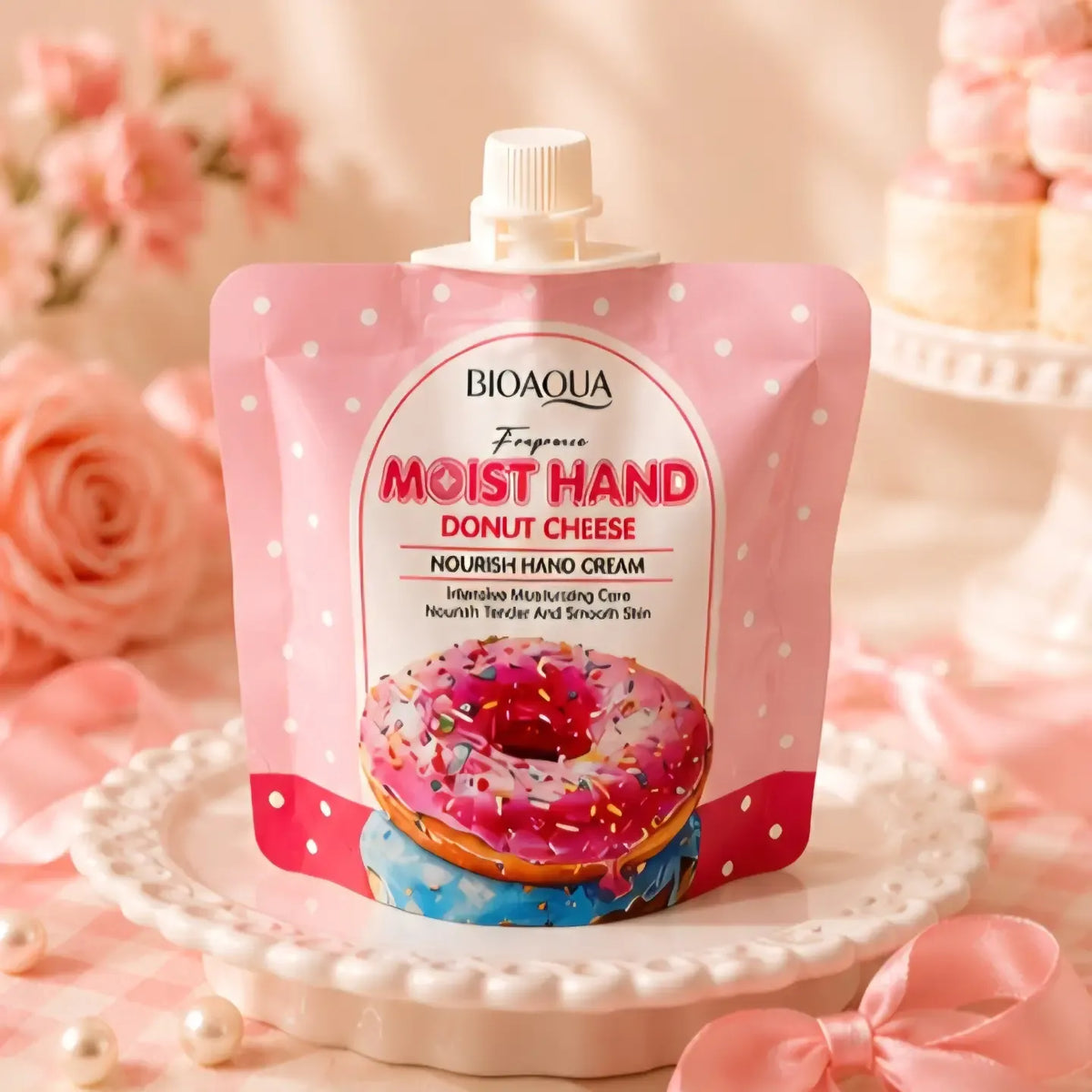 BIOAQUA Moist Hand Donut Cheese Nourish Hand Cream in a pink stand-up squeeze pouch with a donut illustration, displayed on a white plate with pink roses and pastel decor in the background.