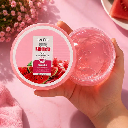 A hand holds an open jar of Sadoer Watermelon Hyaluhydrating Gel alongside its lid, which features a pink and white label with watermelon graphics. The background is a soft pink marble surface decorated with pink flowers and watermelon slices.