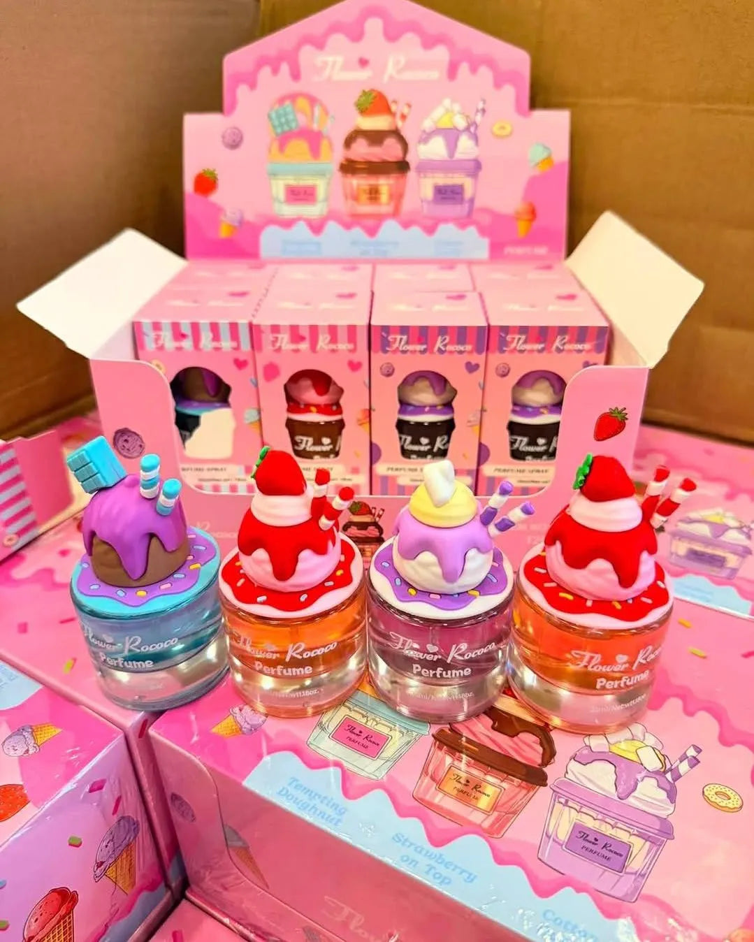 Four small perfume bottles shaped like colorful ice cream desserts are displayed in front of pink retail boxes. The bottles have clear glass bodies with pink, purple, orange, and red liquid, topped with decorative swirls and fruit-like strawberries.