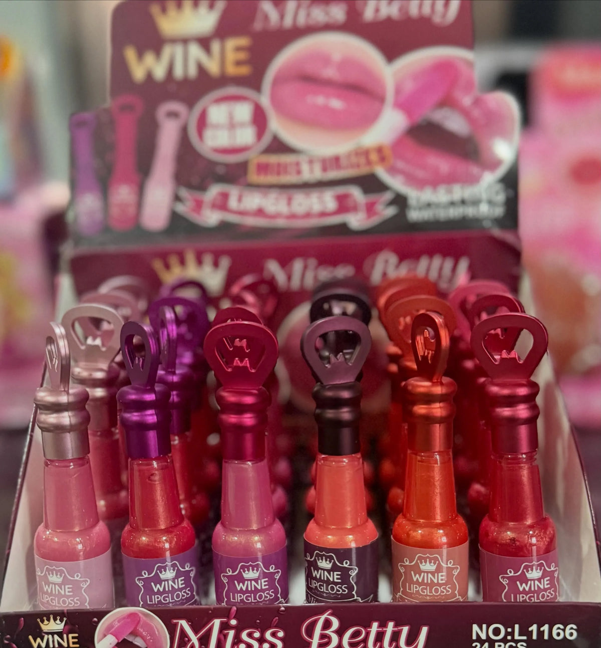 A display box filled with small lip gloss bottles shaped like wine bottles, labeled Miss Betty Wine Lipgloss, in assorted pink, red, orange, and purple shades.