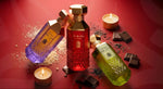 Three luxury perfume bottles with gold caps arranged on a red surface with candles, chocolate pieces, and scattered powder. The bottles contain purple, red, and green liquids with gold labels.
