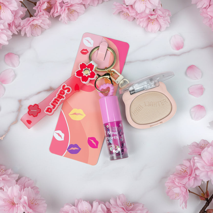 A makeup keychain set featuring a pink eyeshadow compact, a lip gloss tube with purple glitter, and a pink flower charm attached to a metal key ring, displayed on a colorful pink and orange backing card against a black textured background.