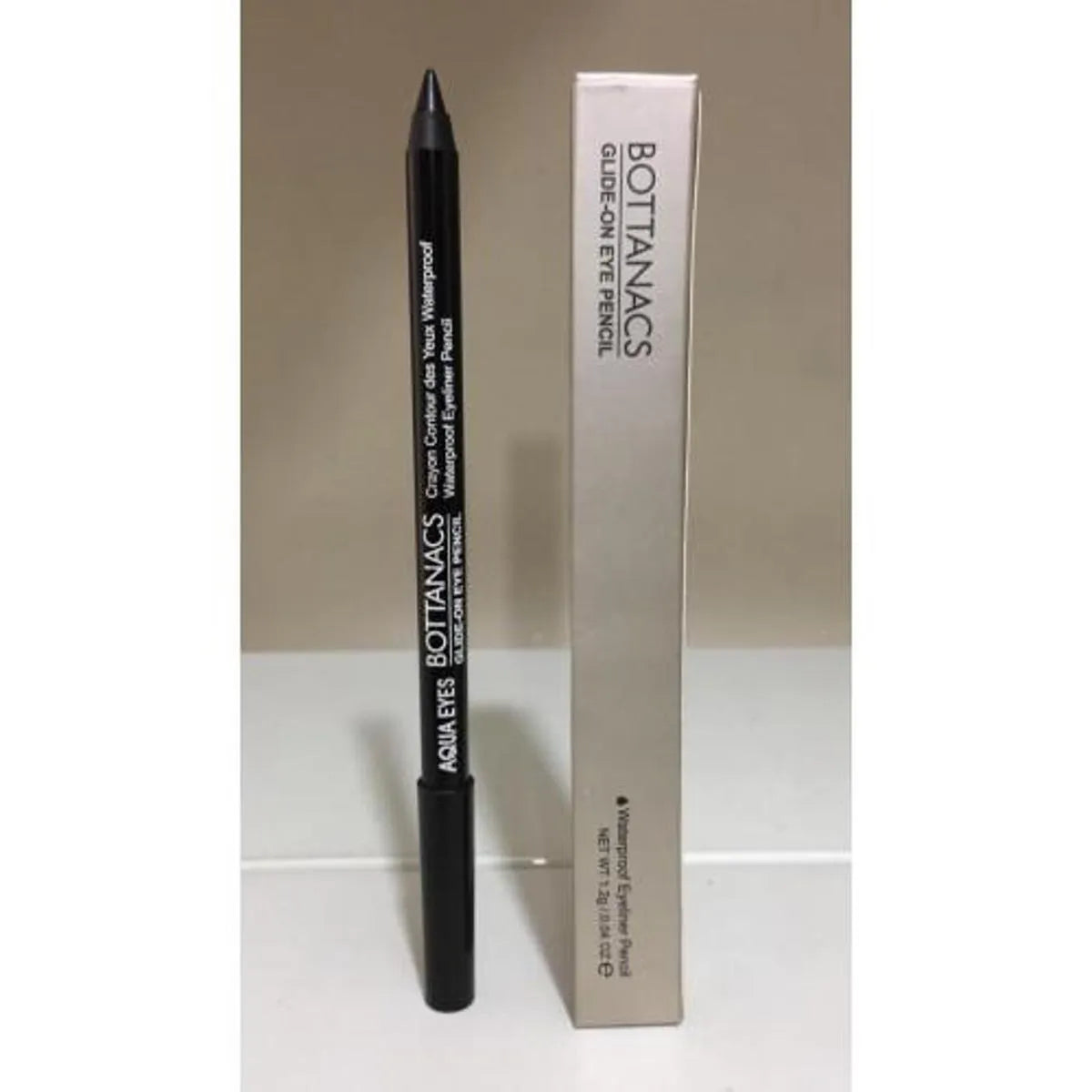 Bottanacs Aqua Eyes Glidden Eye Pencil in black, shown standing next to its silver and white cardboard packaging box on a light surface against a neutral beige background.