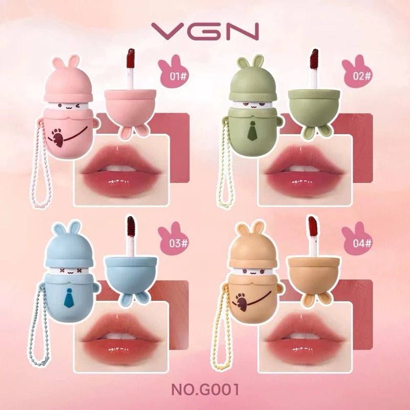 Four cute lip gloss tubes shaped like bunny characters in pink, green, blue, and beige, each shown with a lip applicator and a lip color sample on a soft pink background. The brand VGN is displayed at the top.
