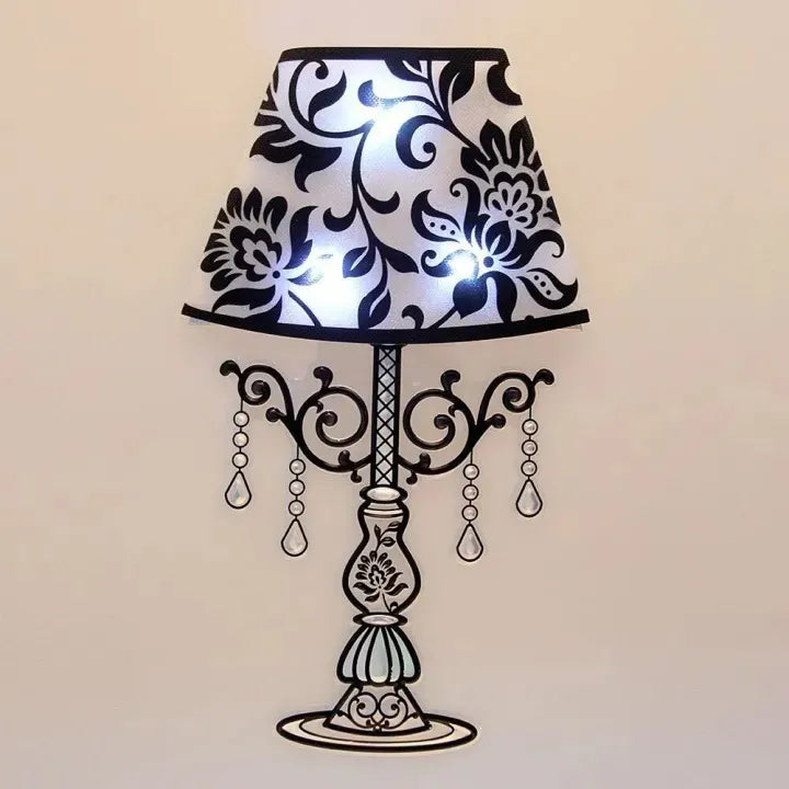 A decorative table lamp with a black and white floral patterned shade, an ornate black metal stand, and hanging teardrop crystal-like accents, shown against a plain light beige wall.