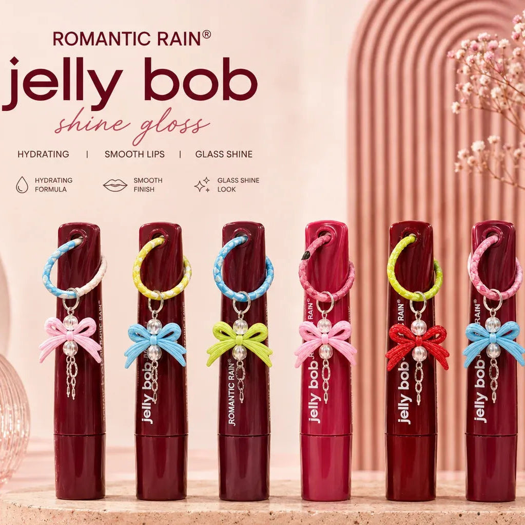 Six tubes of Jelly Bob Shine Gloss lip gloss in assorted shades are lined up on a marble surface with decorative pink props. The tubes feature colorful ribbon bows (pink, blue, yellow, green, red, and light blue) and small pearl charms.