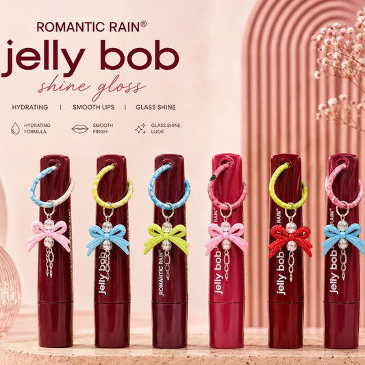 Six tubes of Jelly Bob Shine Gloss lip gloss in assorted shades are lined up on a marble surface with decorative pink props. The tubes feature colorful ribbon bows (pink, blue, yellow, green, red, and light blue) and small pearl charms.