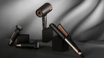 A set of premium hair and facial grooming tools in a dark metallic finish with rose gold accents, displayed on black blocks against a draped gray background. The set includes a hair dryer, a trimmer, a straight razor, and a hair straightener.