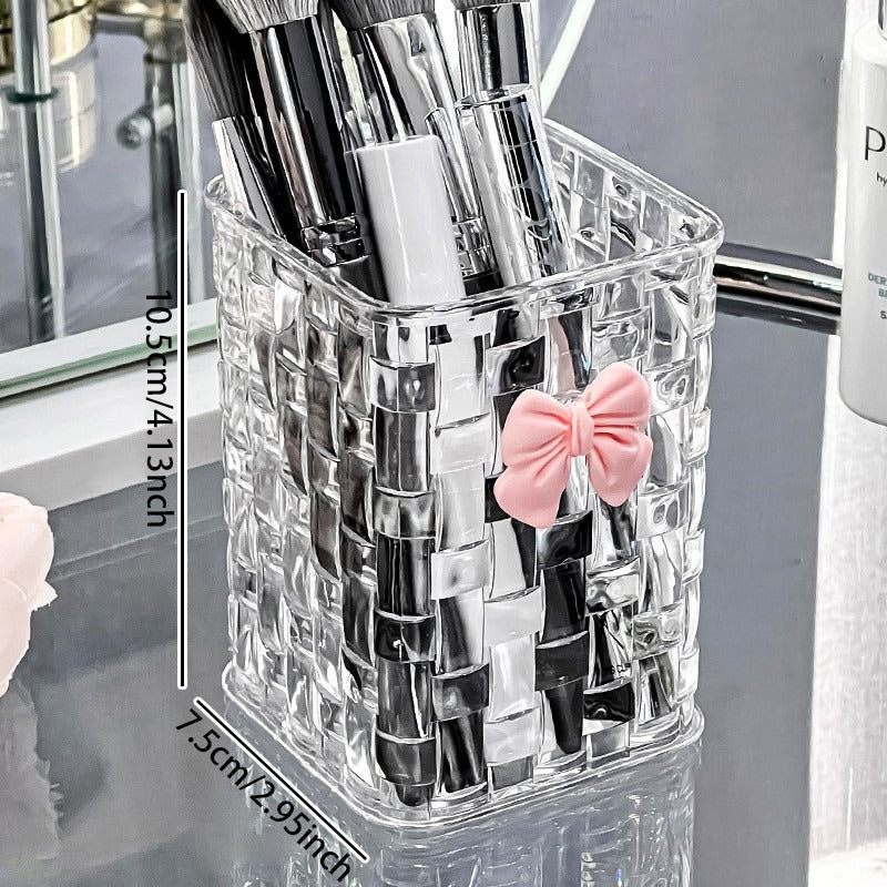 Clear makeup brush holder with lipsticks and a pink bow on a reflective surface.