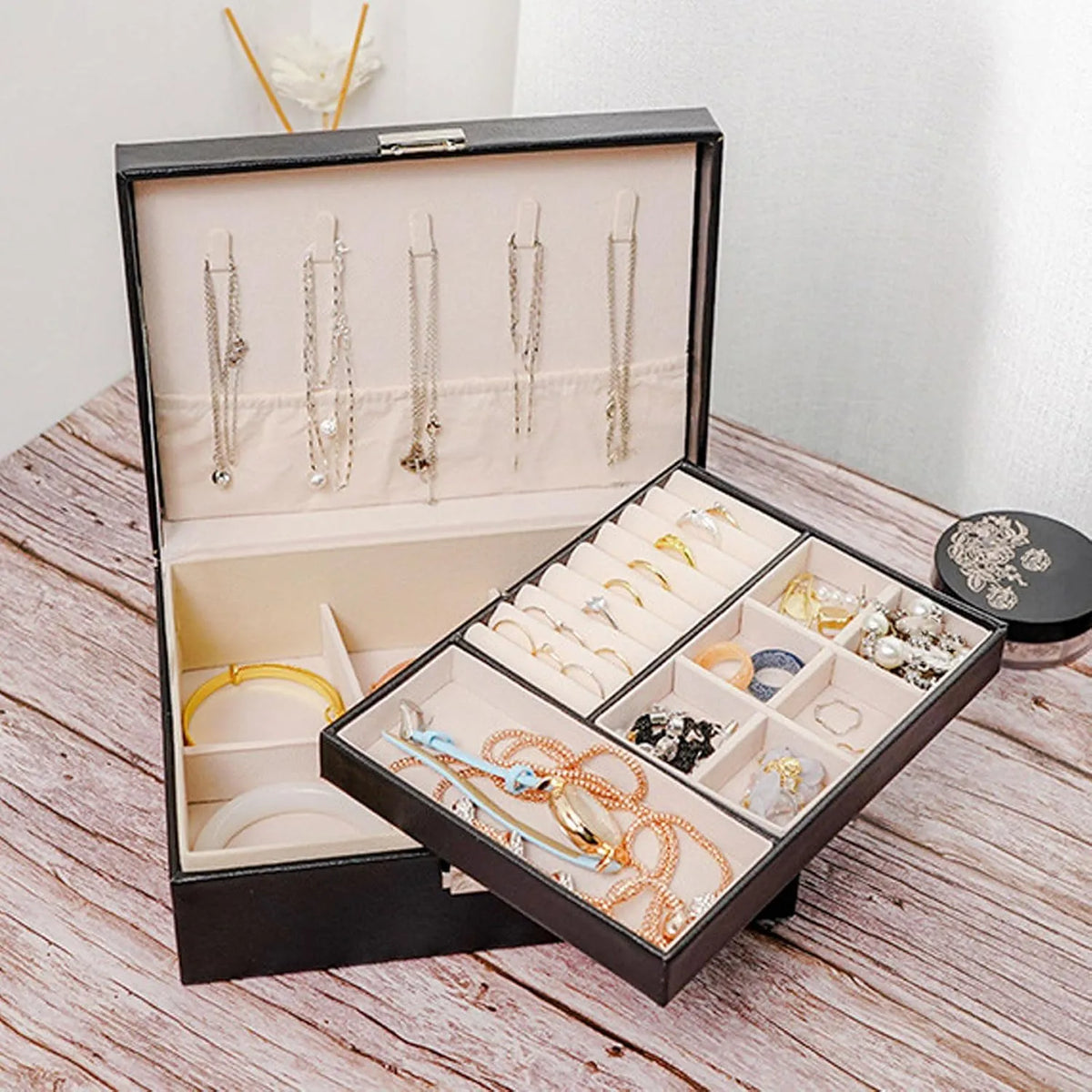 An open black jewelry box with a cream velvet interior, shown on a rustic wooden surface. The box has hanging loops on the inside lid for necklaces, a ring tray, and multiple compartment sections holding rings, earrings, and bracelets.