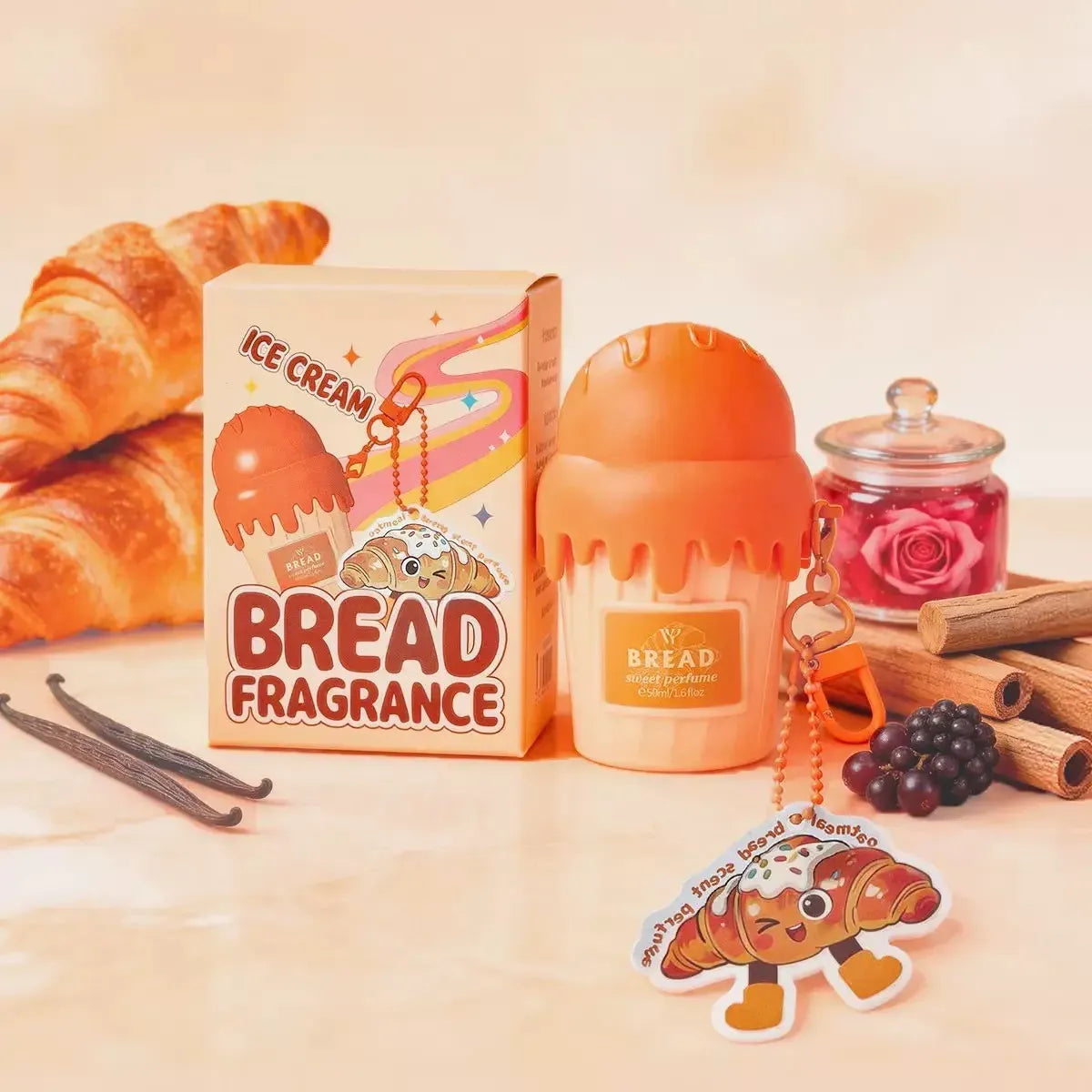 Bread fragrance set with a small orange bread-shaped diffuser, matching product box labeled 'Bread Fragrance' and 'Ice Cream', plus a croissant-shaped keychain charm, styled with croissants, cinnamon sticks, vanilla beans, and a jar of rose petals on a warm beige surface.
