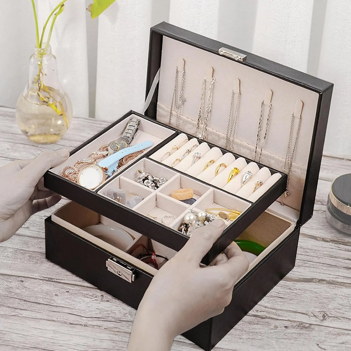 A black jewelry organizer box with a hinged lid, shown open with a person's hands holding the drawer. The organizer contains compartments, necklace stands, and rings, holding various jewelry pieces.
