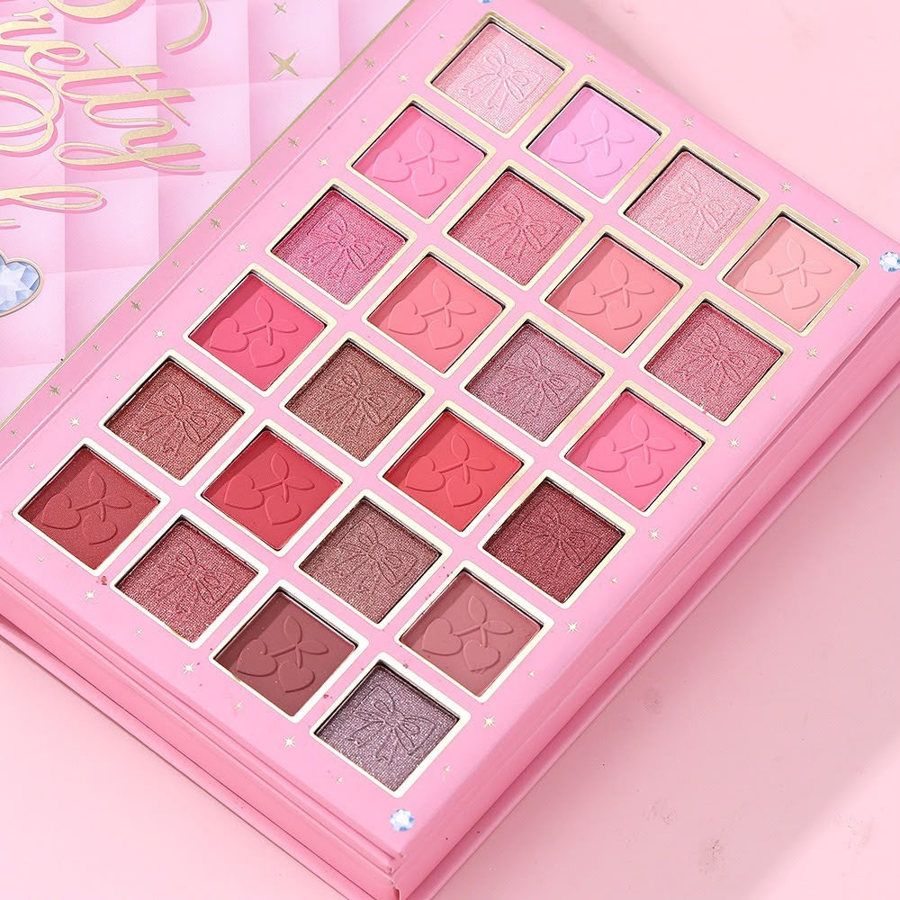 A pink makeup palette with 20 square eyeshadow pans in shades of pink, red, rose gold, and mauve, arranged in a grid. The palette has a glossy pink case with decorative gold and white details, and a matching pink box is partially visible in the background.