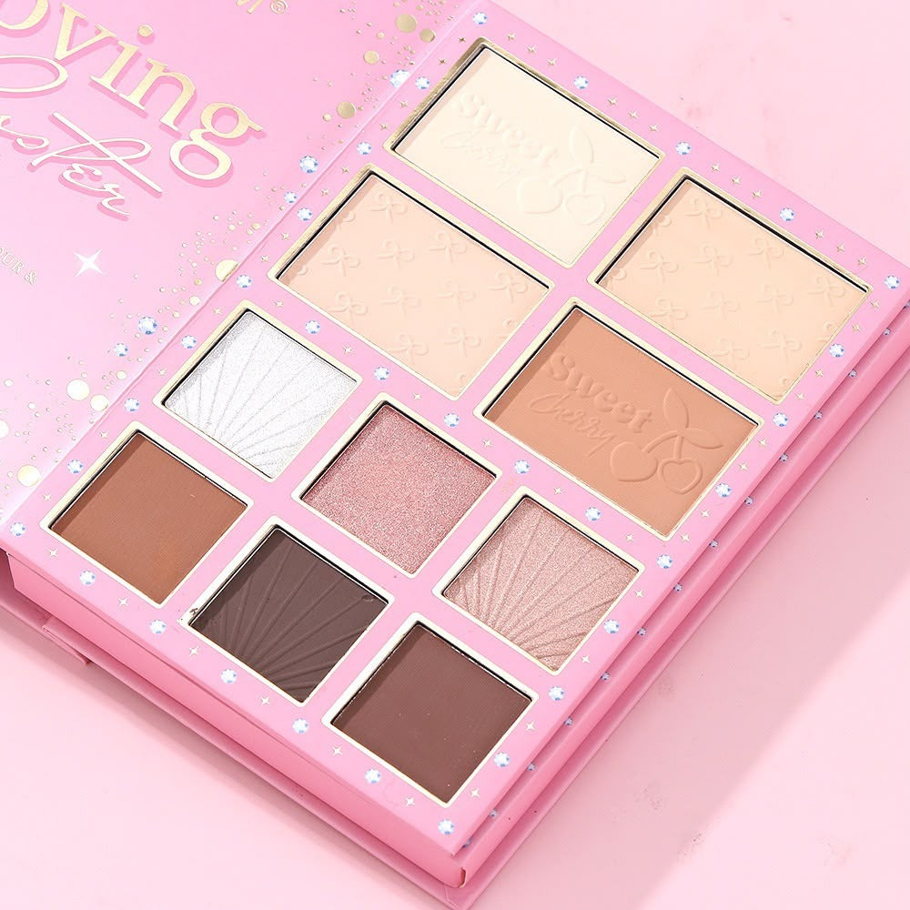 A pink makeup palette with eight square pans of cream and powder products, shown open on a matching pink background. The palette has decorative silver rhinestone accents and partially visible gold lettering on the cover.