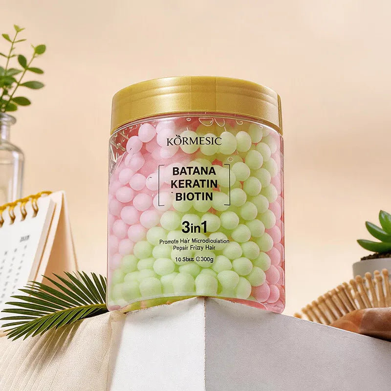 A clear plastic jar of Körmesic Batana Keratin Biotin 3-in-1 hair treatment, filled with pink and light green gel beads and topped with a gold lid, styled on a beige surface with decorative props.