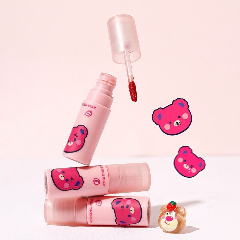Three pink lip gloss tubes with cartoon bear faces are arranged on a pale pink background, with one tube open and its applicator floating above. Two bear-face stickers and a small decorative character accessory sit beside the products.
