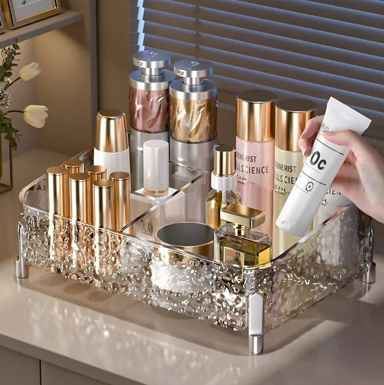 A clear acrylic cosmetic organizer tray with a glittery textured base, shown on a bathroom or vanity counter holding various skincare bottles and tubes. A hand is holding a white tube, and the background features gray window blinds with text overlays describing the product's large mouth design for easy access.
