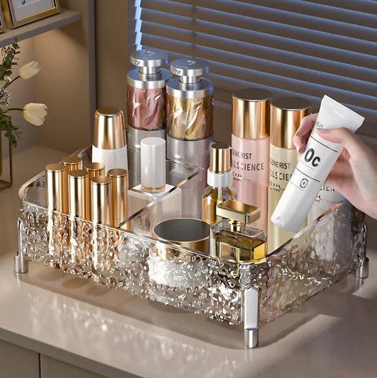 A clear acrylic cosmetic organizer tray with a glittery textured base, shown on a bathroom or vanity counter holding various skincare bottles and tubes. A hand is holding a white tube, and the background features gray window blinds with text overlays describing the product's large mouth design for easy access.