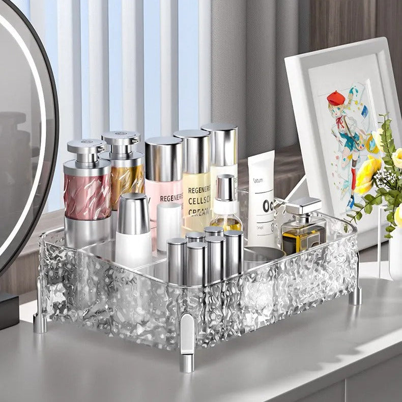 A clear acrylic makeup organizer tray with a textured, rhinestone-like pattern and small legs, holding assorted cosmetic bottles and tubes. The tray sits on a white vanity table in a bright room with vertical blinds, a mirror edge visible on the left, and a framed print on the right
