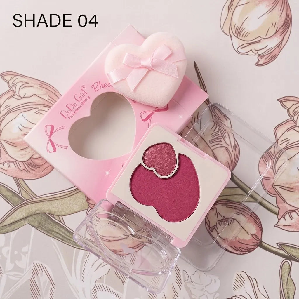 A heart-shaped pink eyeshadow palette shown open with a matching heart-shaped sponge and a small heart-shaped makeup brush, on a floral background. The text 'SHADE 04' appears in the top left corner.