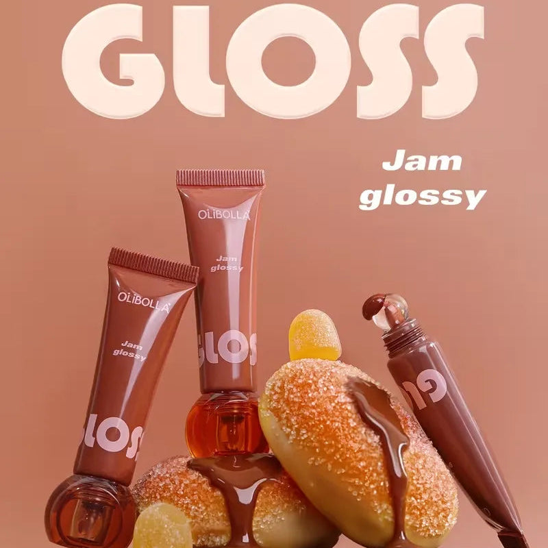 Three Gloss Jam glossy tubes arranged on a wooden pedestal with a glazed donut, jam pieces, and a chocolate splash at the bottom. Large text at the top reads 'GLOSS' and smaller text reads 'Jam glossy'.