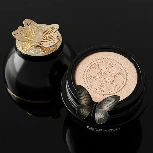 Close-up with butterfly: “Gegemoon cream foundation cushion beside a black jar with gold butterfly lid”