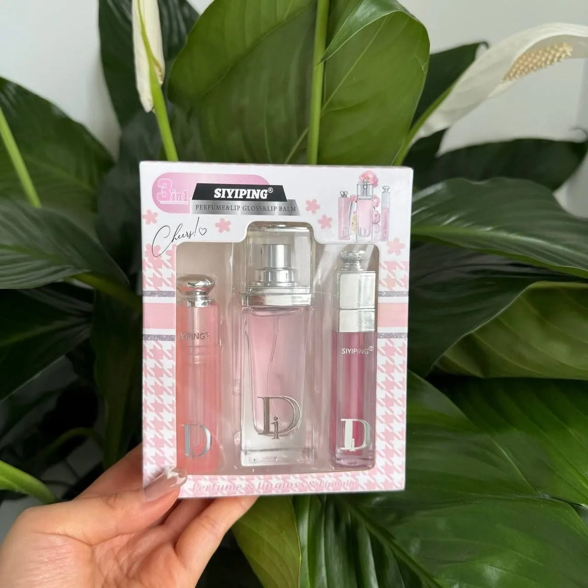 A hand holds a boxed fragrance gift set in front of large green plant leaves. The package contains three pink-toned perfume/cologne bottles, including a central clear bottle with a silver cap and a visible Dior logo.