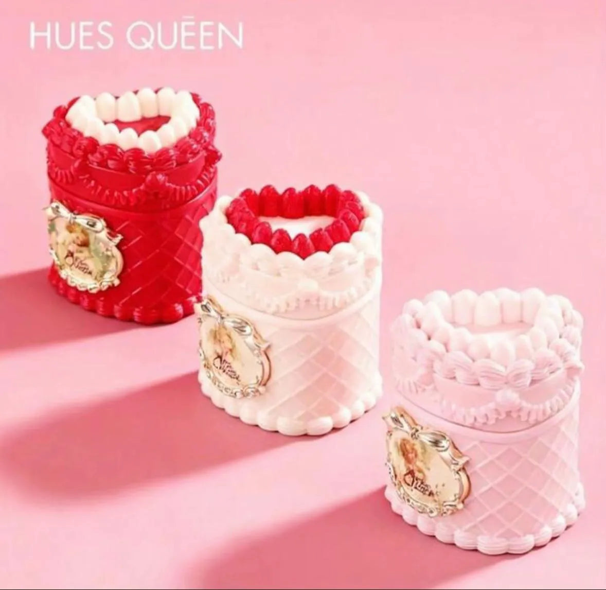 Three decorative cylindrical containers from HUES QUEEN in red and white, and pale pink, arranged on a solid pink background. Each container features a quilted pattern, a ruffled trim, a small bow, and an ornate gold crest with a portrait.