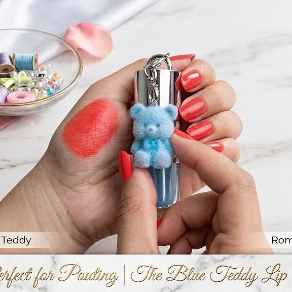 A close-up of hands holding a lip tint tube with a small blue teddy bear applicator. The background is a white marble surface with pink petals, a small bowl of colorful thread spools, and a gold key.