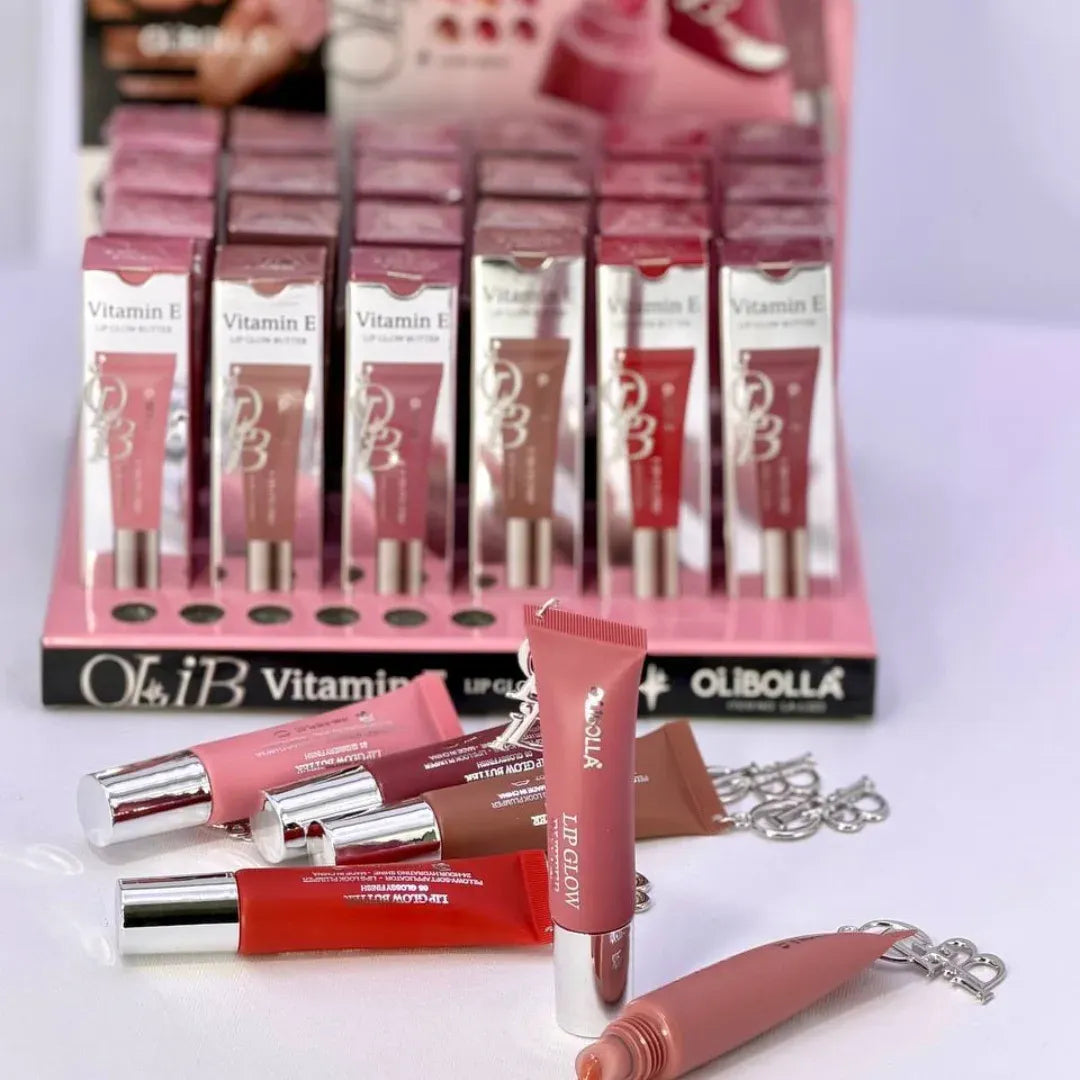 A display of Olibolla Vitamin E lip gloss tubes in various shades, including a pink display box and several individual tubes with silver caps arranged in front on a light surface.