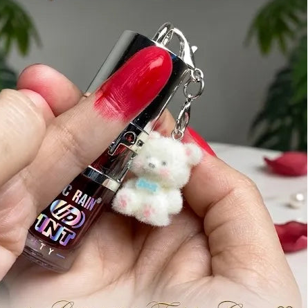 A hand holds a small cosmetic tube labeled THE G RAIN TINT Collection with a white teddy bear keychain charm. The scene is styled with red nail polish, rose petals, pearls, and a gold key on a marble surface, with green foliage in the background.