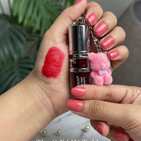 A close-up of hands holding a dark red lipstick tube with a pink teddy bear keychain charm. A swatch of the lipstick color is visible on the back of one hand.