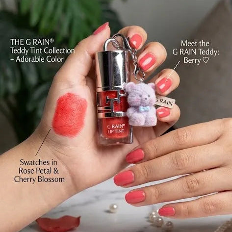 A close-up of hands holding a G.RAIN Jelly Bob Shine Gloss bottle, with coral pink nail polish swatches on the fingers and scattered rose petals and pearls on a marble surface.